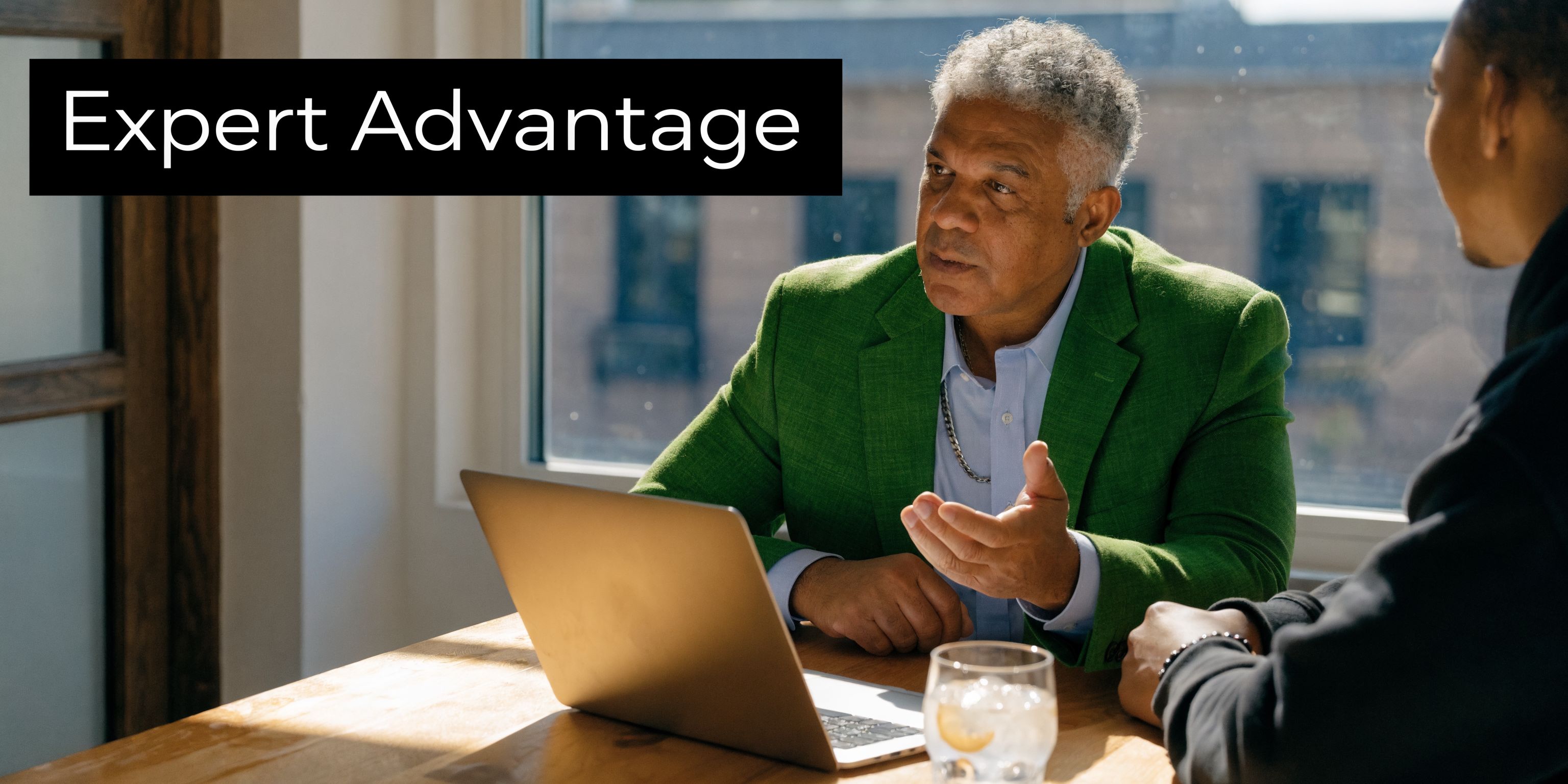 A professional man with gray hair in a green jacket speaking to a client while using a laptop.
